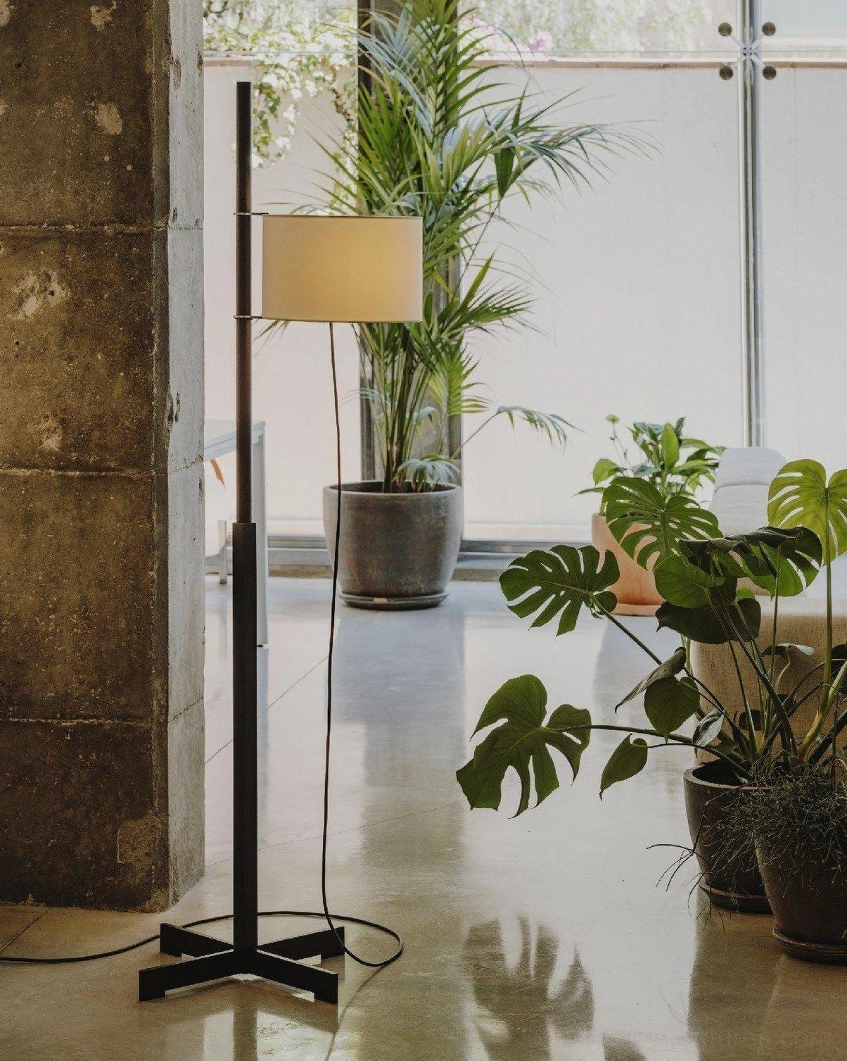 Black metal modern floor lamp with beige fabric shade in industrial sunlit space with plants