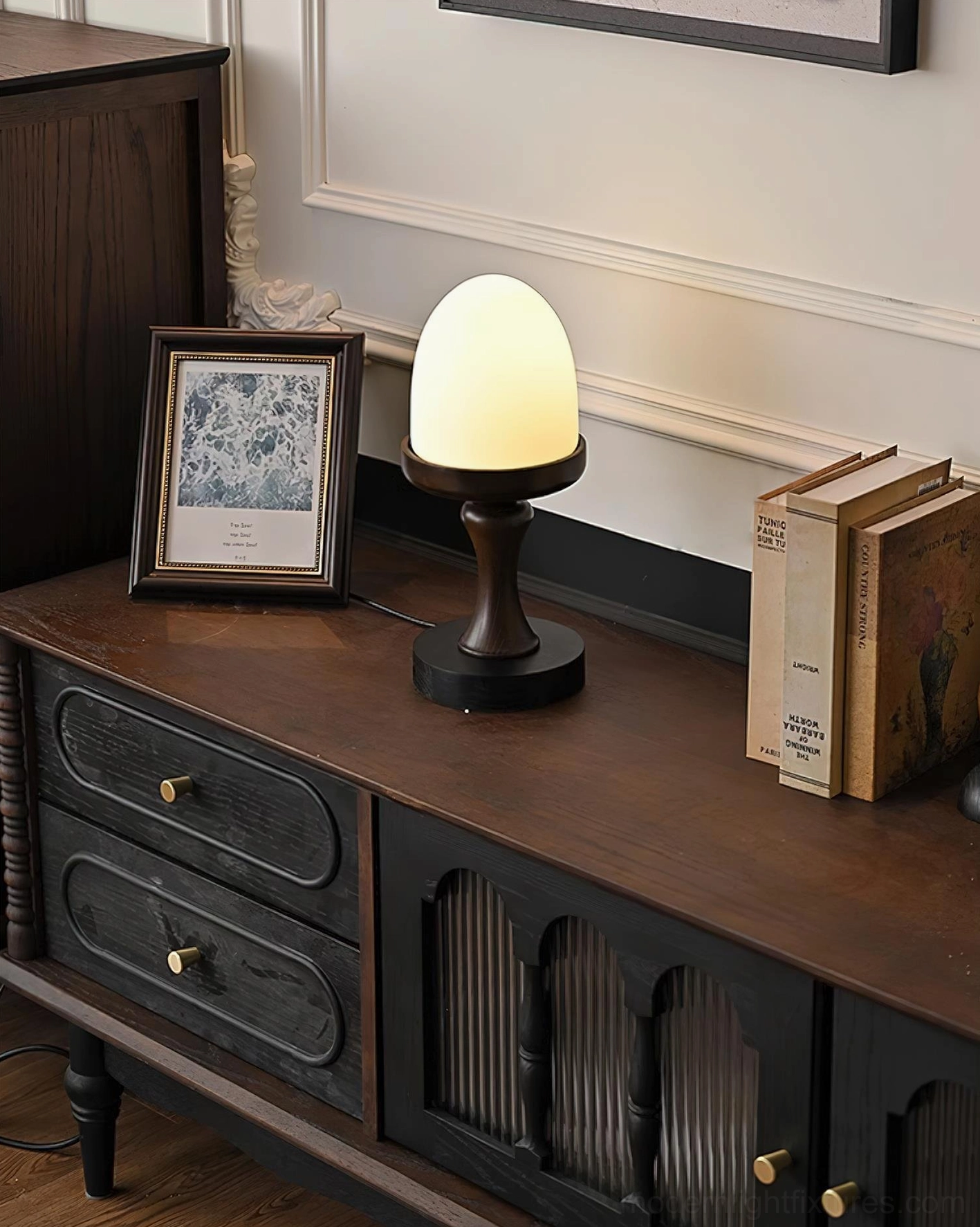 Lit natural wood base lamp with egg-form frosted shade on long retro console with wainscoting wall backdrop