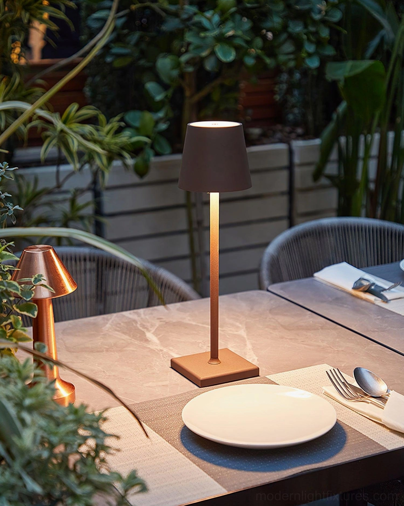 Dark tapered shade bronze metal stem modern table lamp on outdoor dining patio with ambient warm lighting