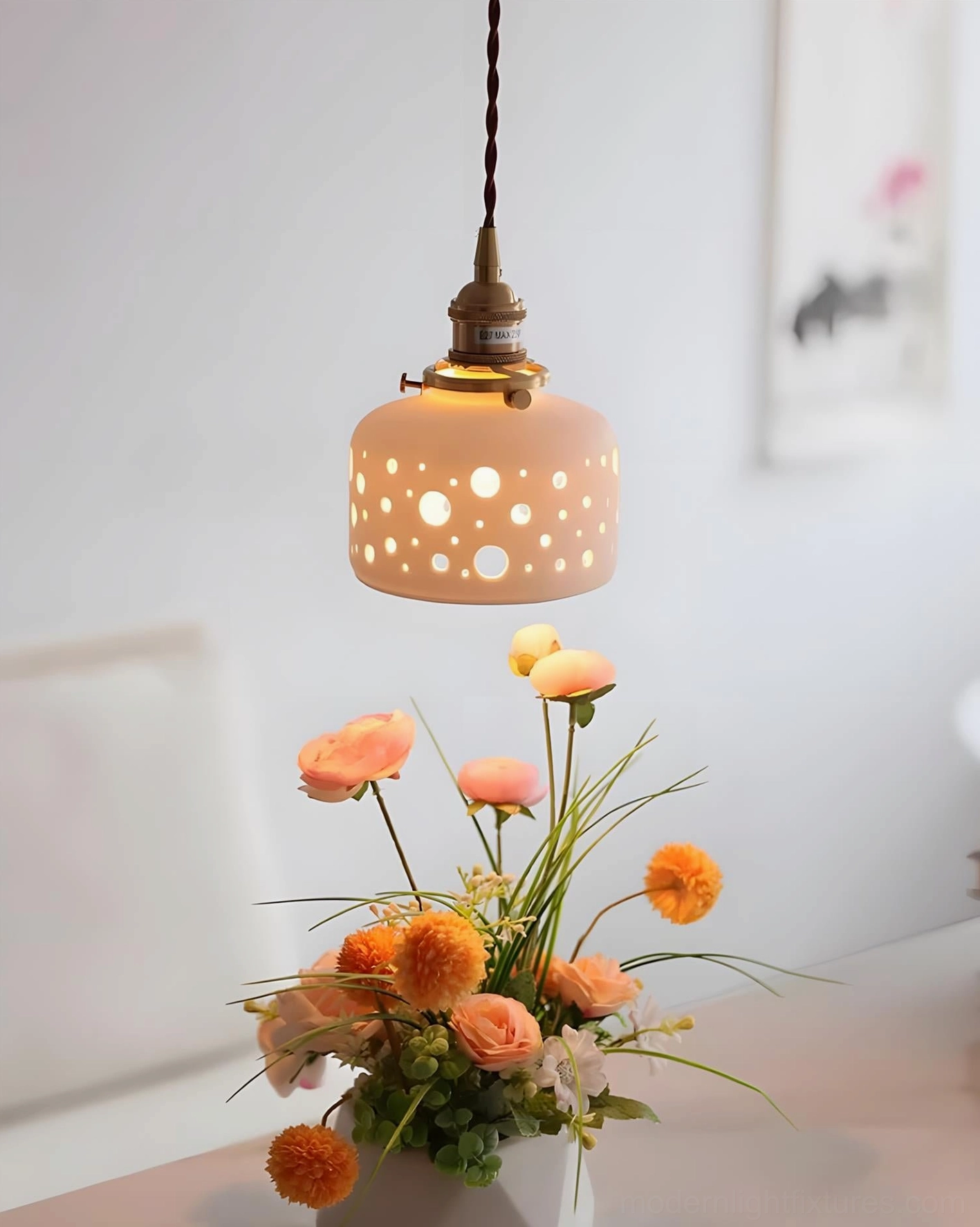 Softly illuminated Moonveil ceramic pendant lamp with perforated shade beside fresh flower arrangement, angled perspective