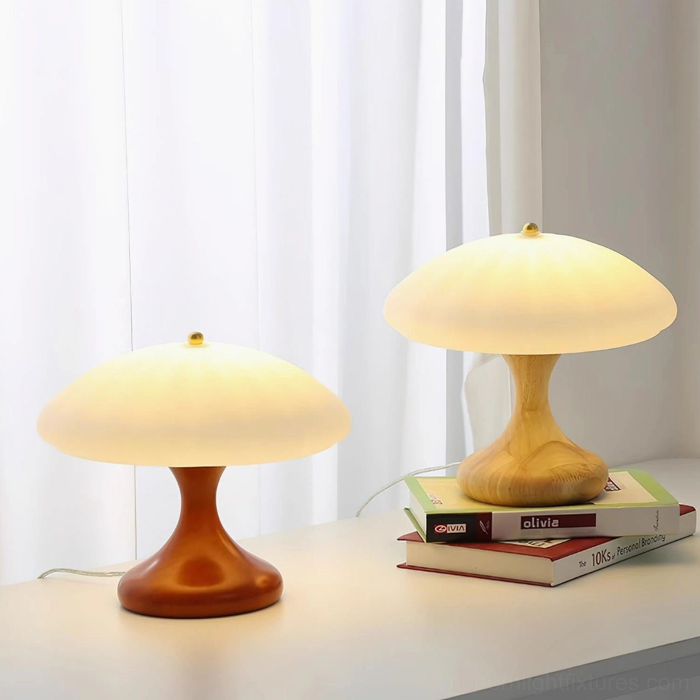 Pair of mushroom table lamps in light and dark wood finishes arranged on stacked books with curtain backdrop, left perspective home styling