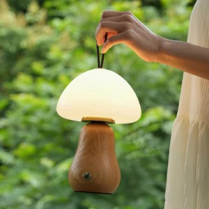 Portable mushroom table lamp held by carrying strap in outdoor garden setting with green foliage background and warm glowing light