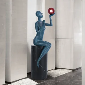 Modern seated sculpture floor lamp with teal figure in minimalist stone lobby with gravel flooring - three-quarter angle view