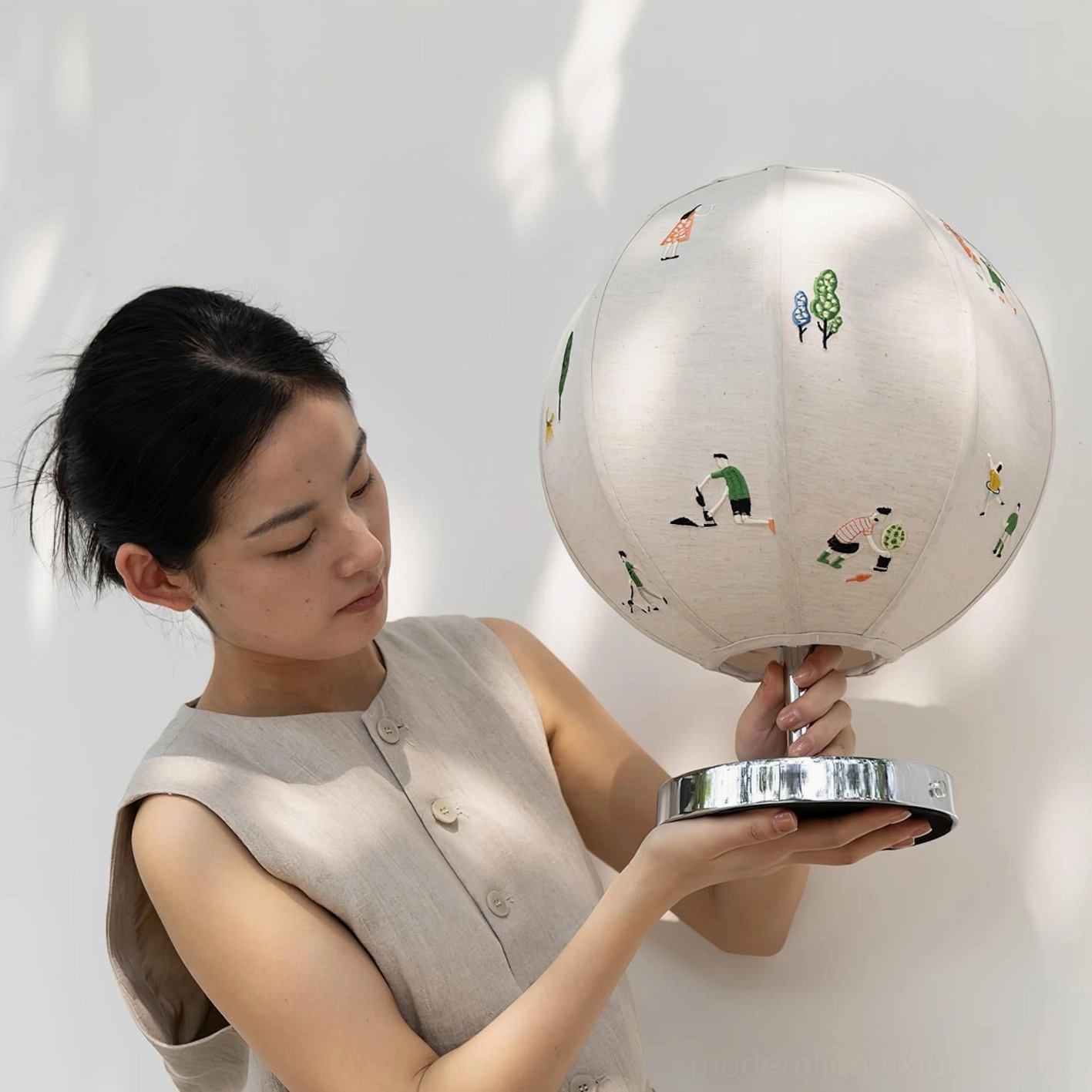 Woman holding modern embroidered fabric table lamp in home setting, side three-quarter angle showing texture of linen shade and chrome base