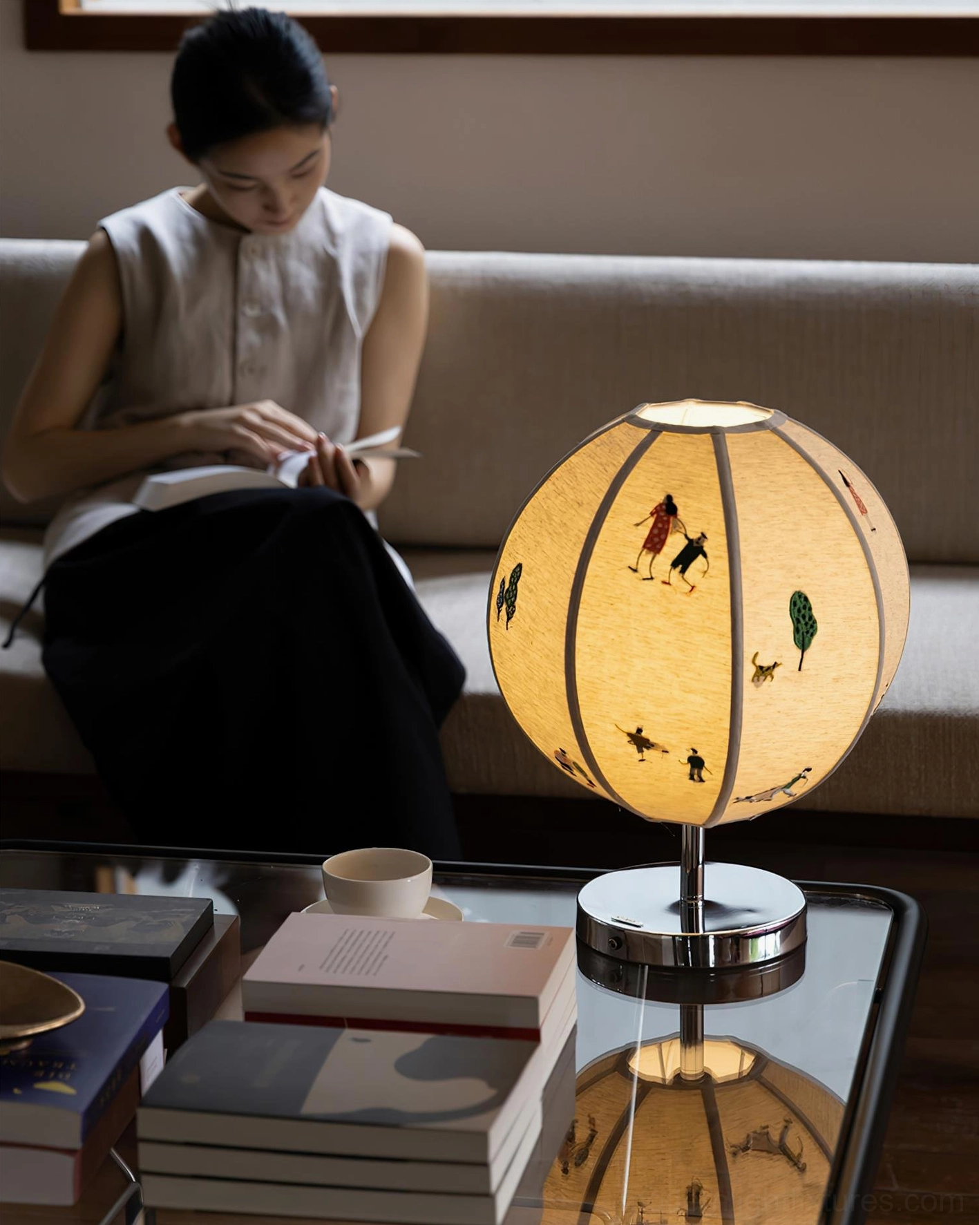 Lit embroidered table lamp on coffee table in home reading area with stacked books and woman reading, warm white light ambiance
