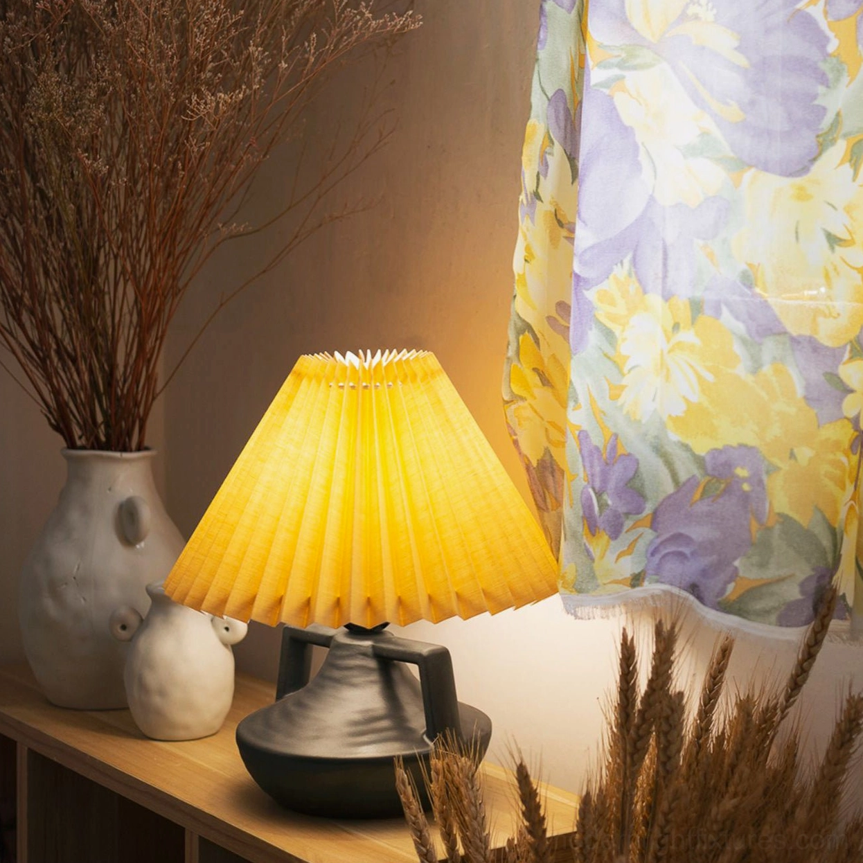 Navia Pleated Table Lamp lit on living room side table with warm yellow shade and dark grey ceramic base
