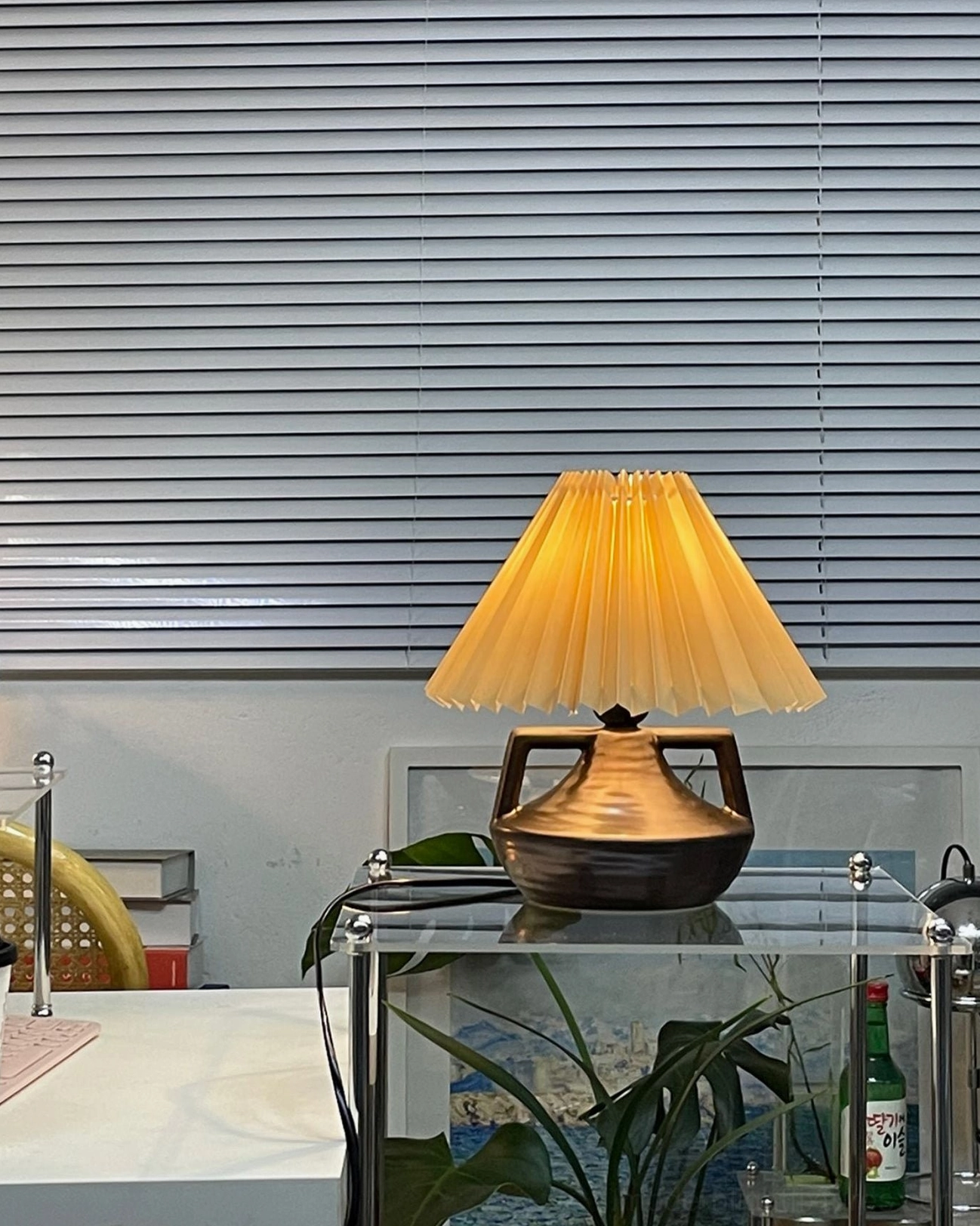 Navia Pleated Table Lamp three-quarter view on modern glass side table with dark brown ceramic base and green plant