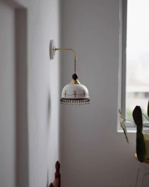 Noemi wall lamp in residential interior, warm ceramic shade with brass arm, indoor side perspective with natural window light