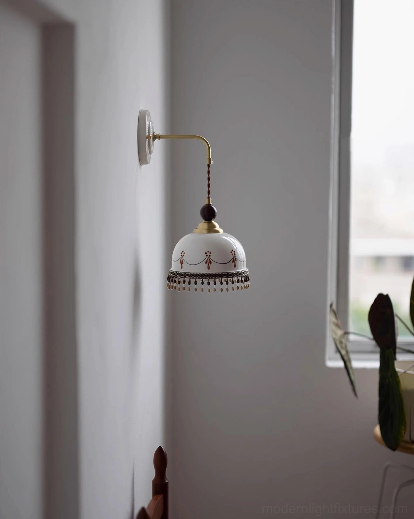 Noemi wall lamp in residential interior, warm ceramic shade with brass arm, indoor side perspective with natural window light