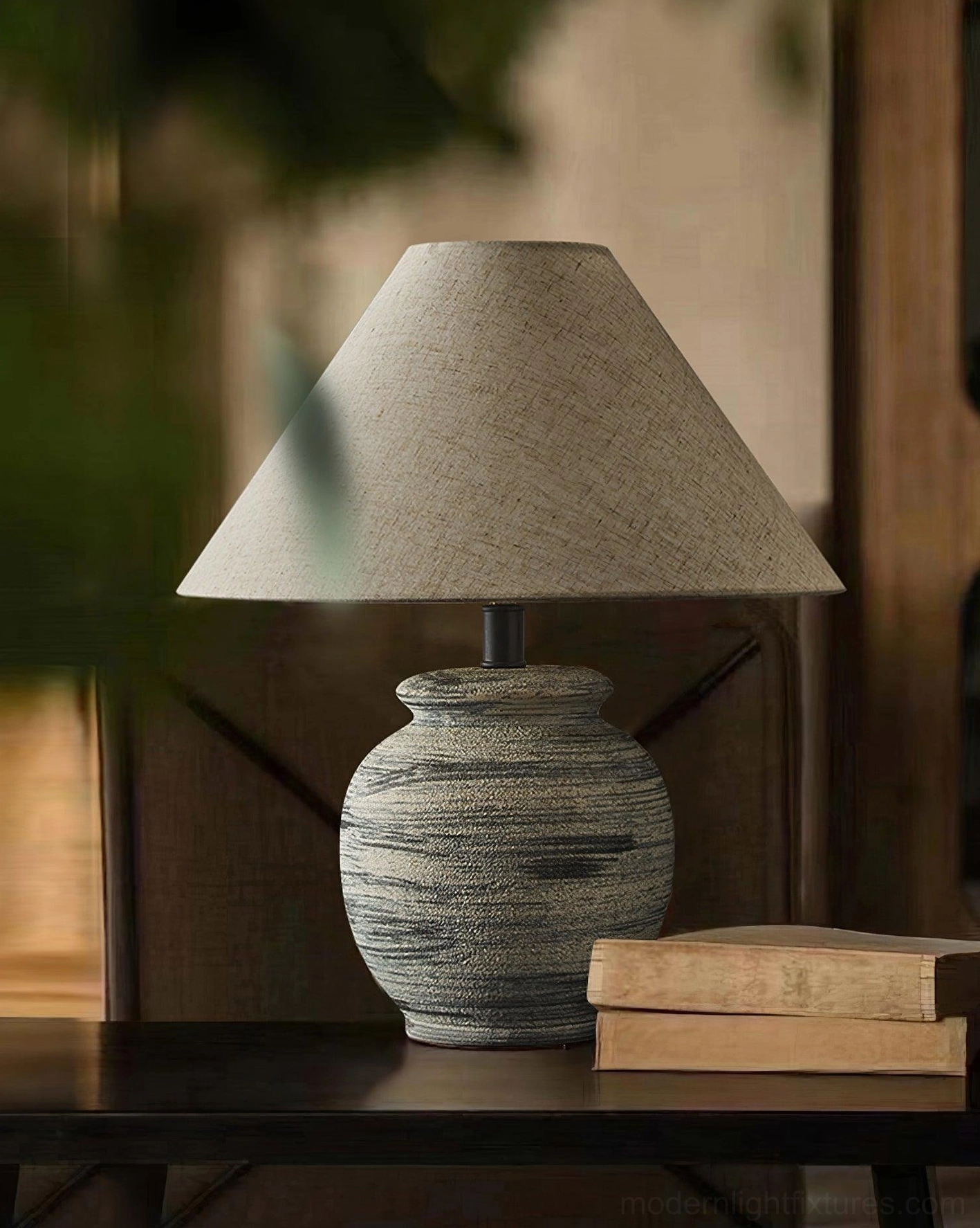 Norma ceramic table lamp side-angle view with stacked books and green foliage background, warm tones