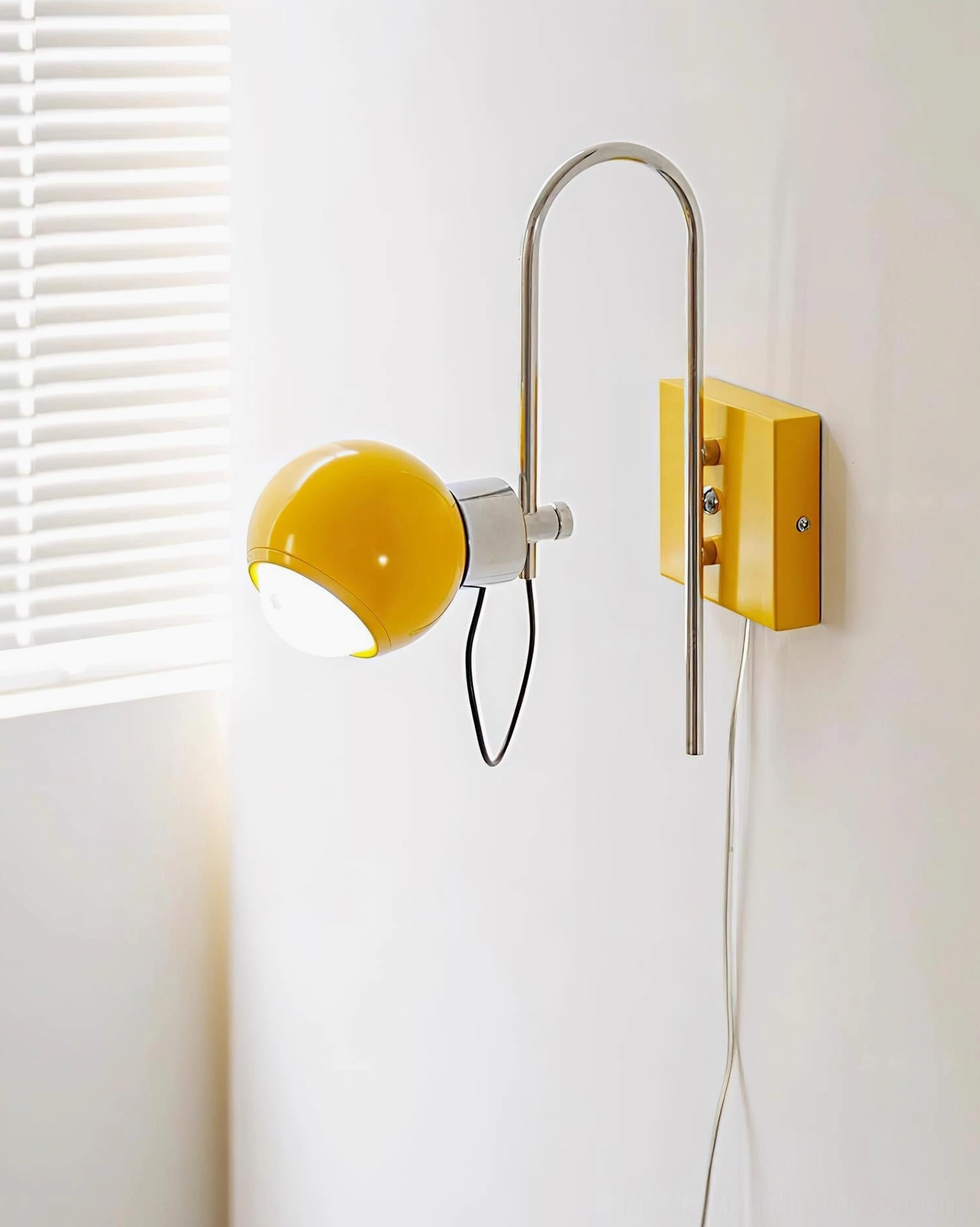 Nova Dot yellow wall sconce with illuminated spherical shade and white exposed power cord against white window blind wall