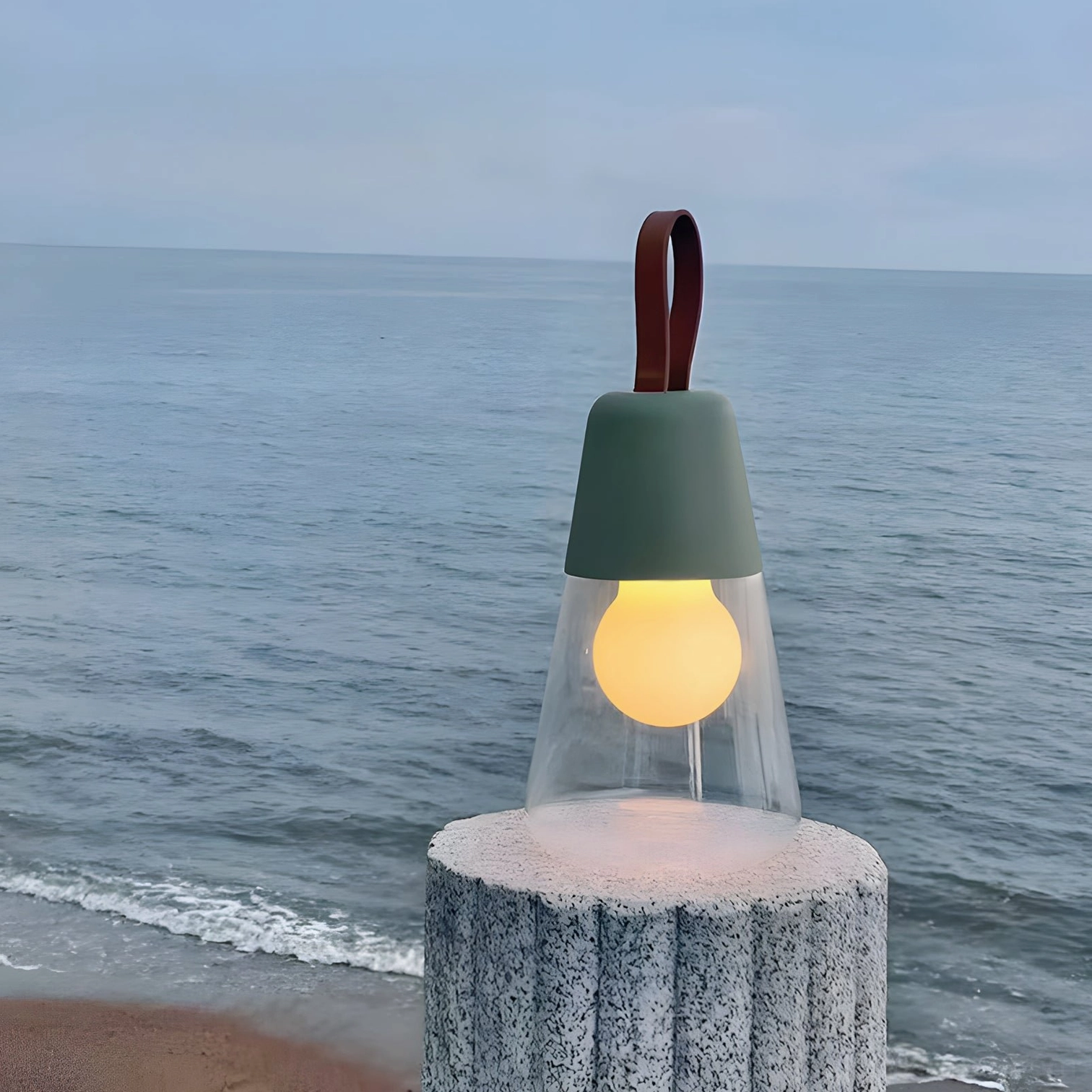 Dark green Oceana portable table lamp with leather strap displayed on coastal beach setting, warm ambient lighting
