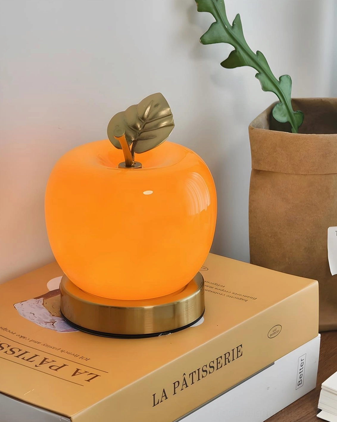 Orange apple lamp three-quarter view on desktop vanity corner next to potted plant and bakery-themed book