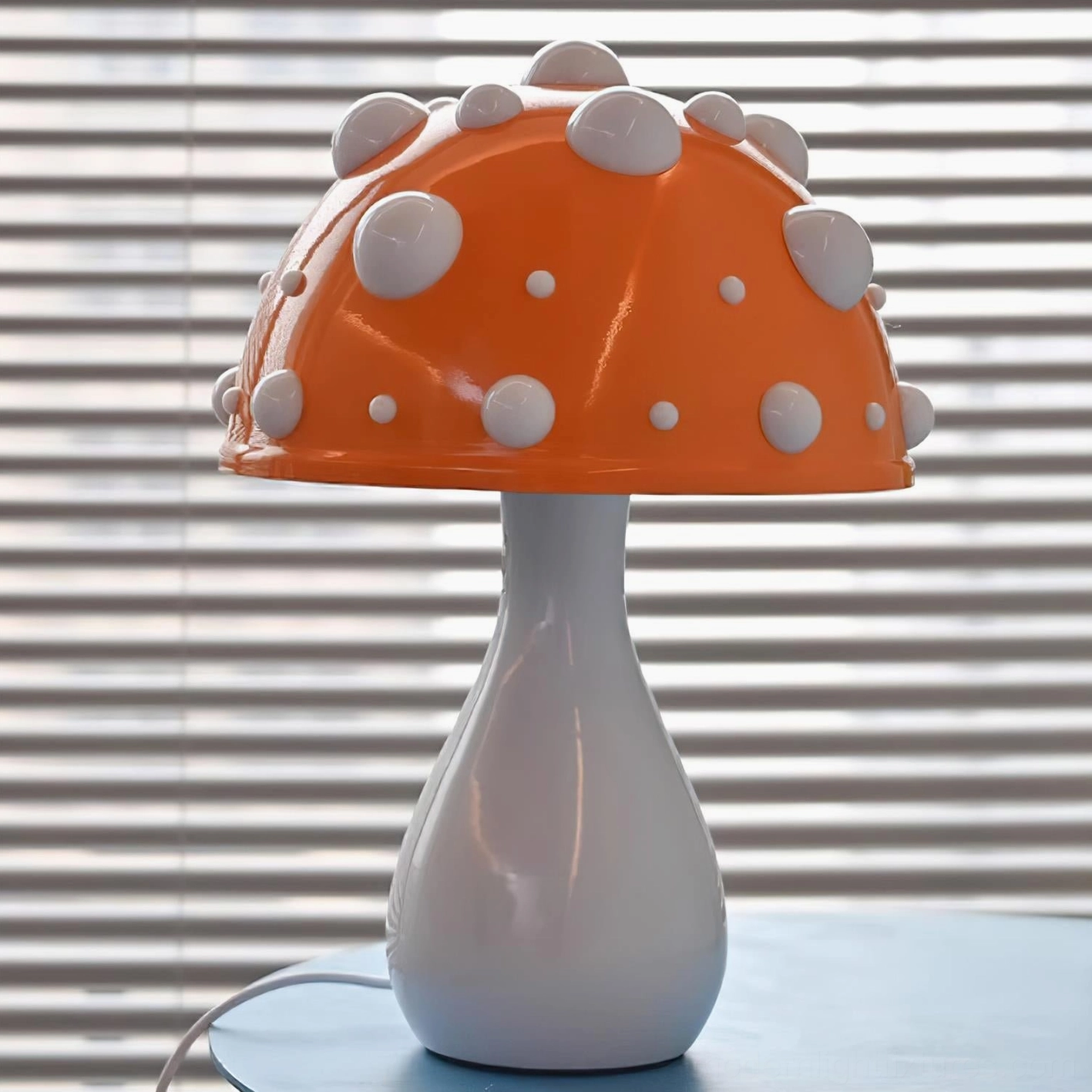 Orange spotted mushroom table lamp with raised white dots on cap, frontal view on desk with window blinds backdrop