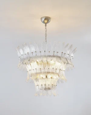 Illuminated palmette chandelier with warm glowing light reflecting through clear glass tiers and metallic frame against off-white wall