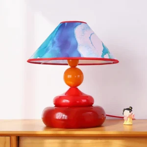 Paloma table lamp on wooden tabletop with red ceramic base, watercolor shade, and decorative figurine in indoor home setting