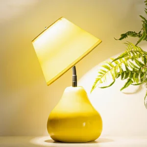 Yellow pear table lamp with lime green fabric shade, front three-quarter view with warm ambient lighting and decorative fern beside it