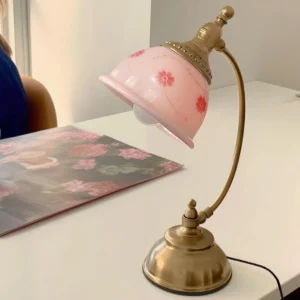 Pink floral ceramic desk lamp with curved brass arm and matching gold base, side three-quarter view in home office