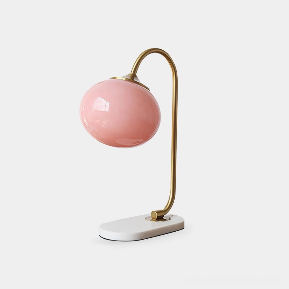 Pink glass globe table lamp with gold metal curved frame and white marble base, front three-quarter view on studio background