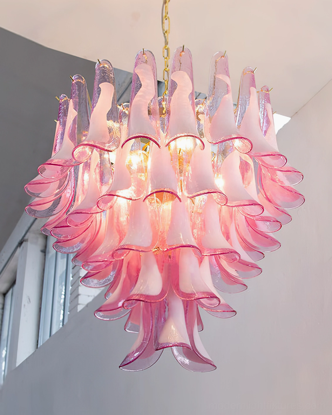 Illuminated pink tulip petal chandelier in residential interior with natural window light, warm glowing ambient lighting effect on tiered floral silhouette