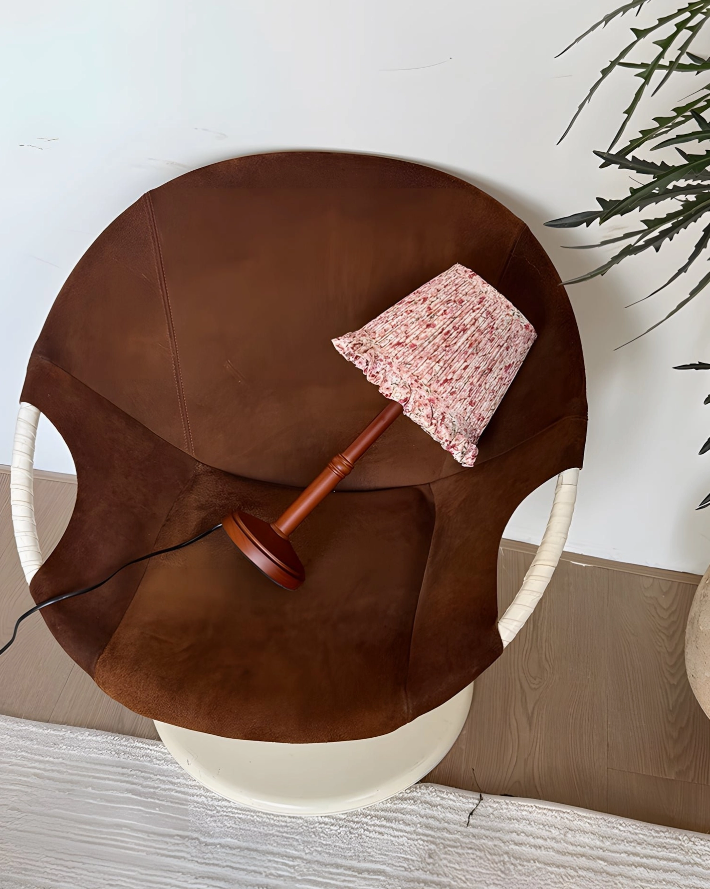 Pleated fabric table lamp on brown leather chair with green houseplant, overhead tilted angle