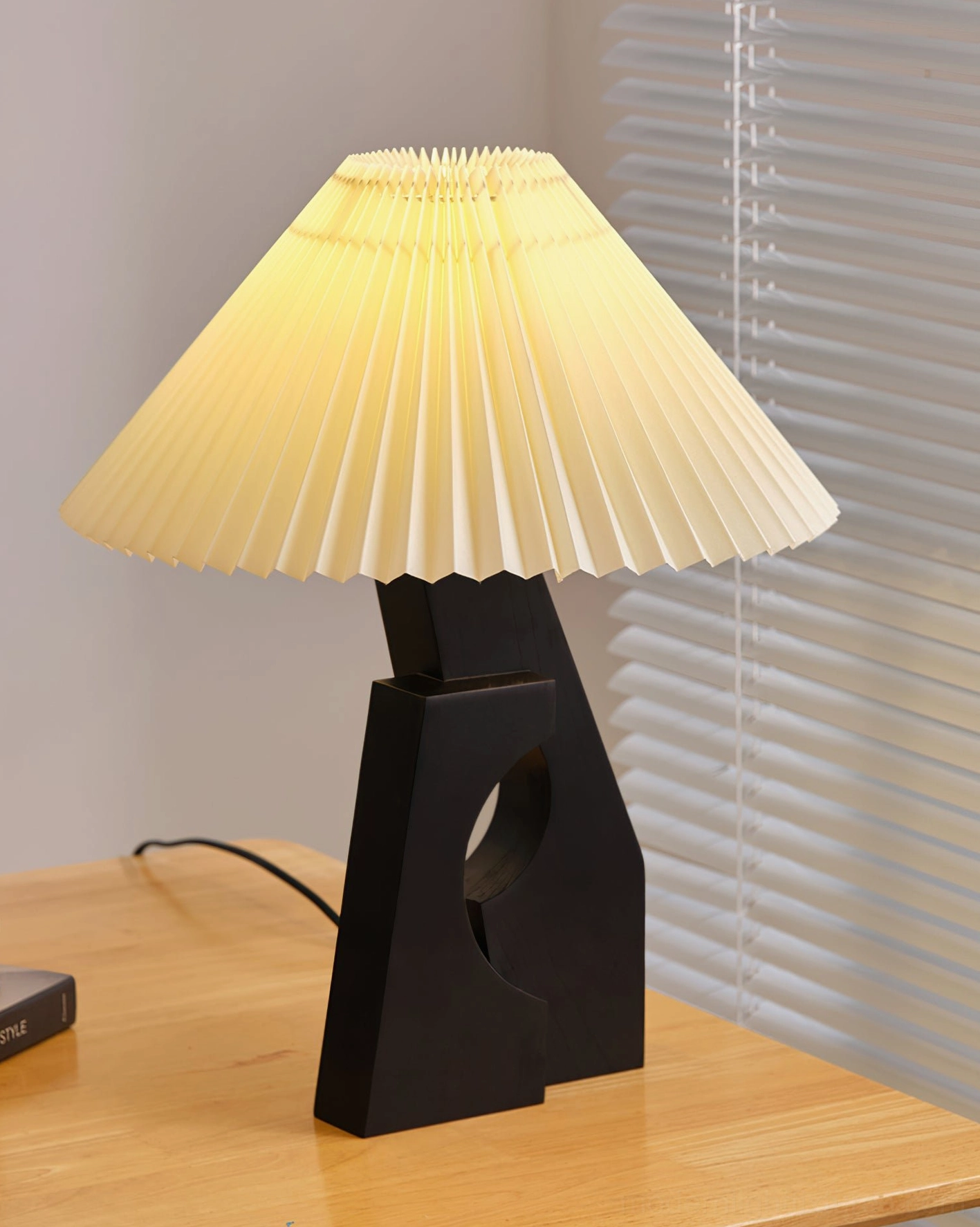 Modern pleated table lamp with black hollow wood base illuminated, warm yellow glow on home office desk with window blinds