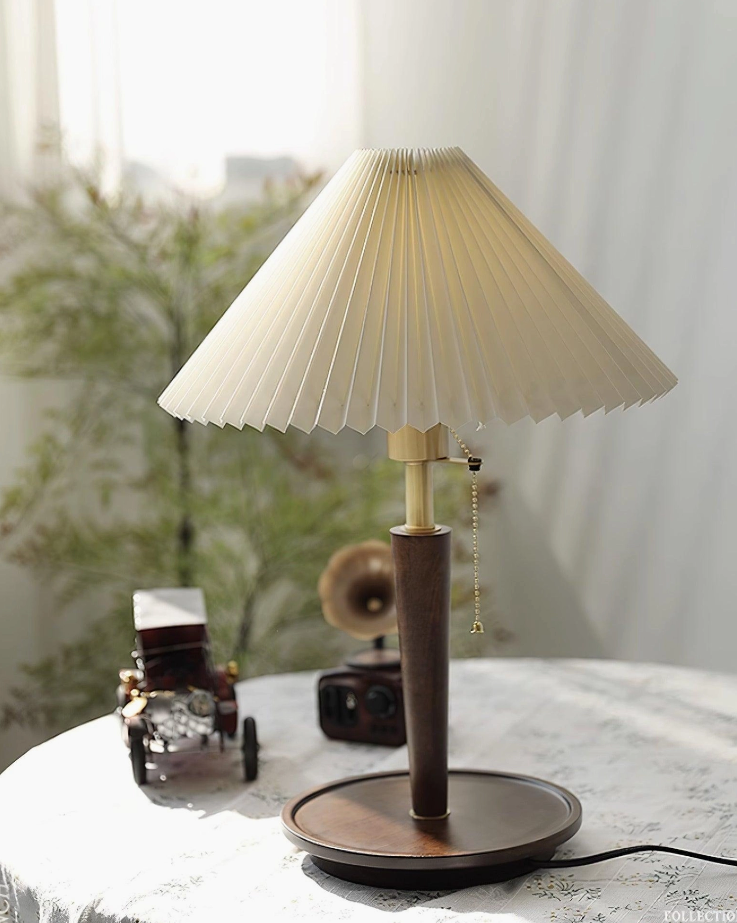 Suki pleated table lamp right side view on round lace table with soft indoor lighting and vintage toy accessories