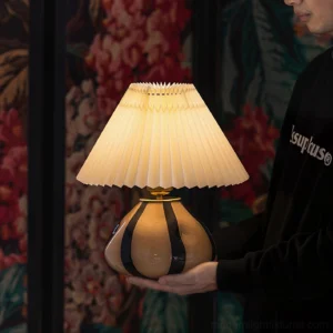 Illuminated pleated table lamp held in hand, warm LED light glowing through textured fabric shade in interior setting