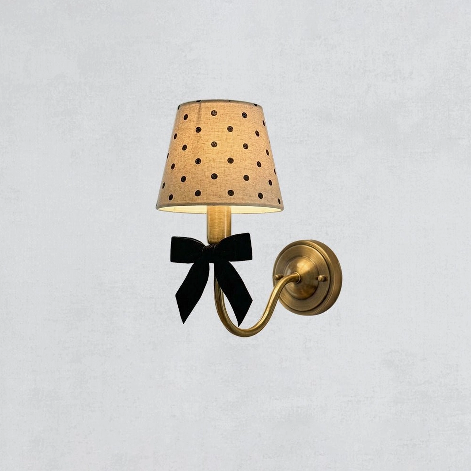 Polka dot wall sconce with brass metal frame and black velvet bow on light gray background, frontal flat studio view showing fabric shade details