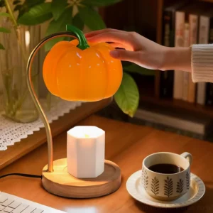 Hand adjusting pumpkin candle warmer lamp on home office desk with coffee cup and potted plant, creating cozy workspace ambiance