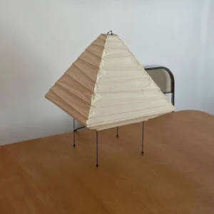 Modern pyramid table lamp side view in bedroom setting, warm light glowing from folded paper shade