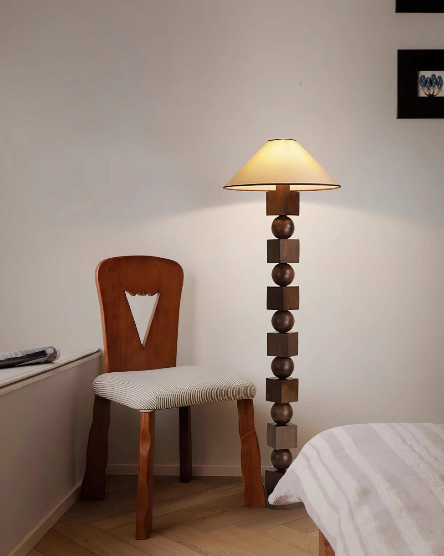 Mid-century modern sculptural wooden floor lamp providing warm ambient light in bedroom corner with wooden chair and bed backdrop, eye-level view