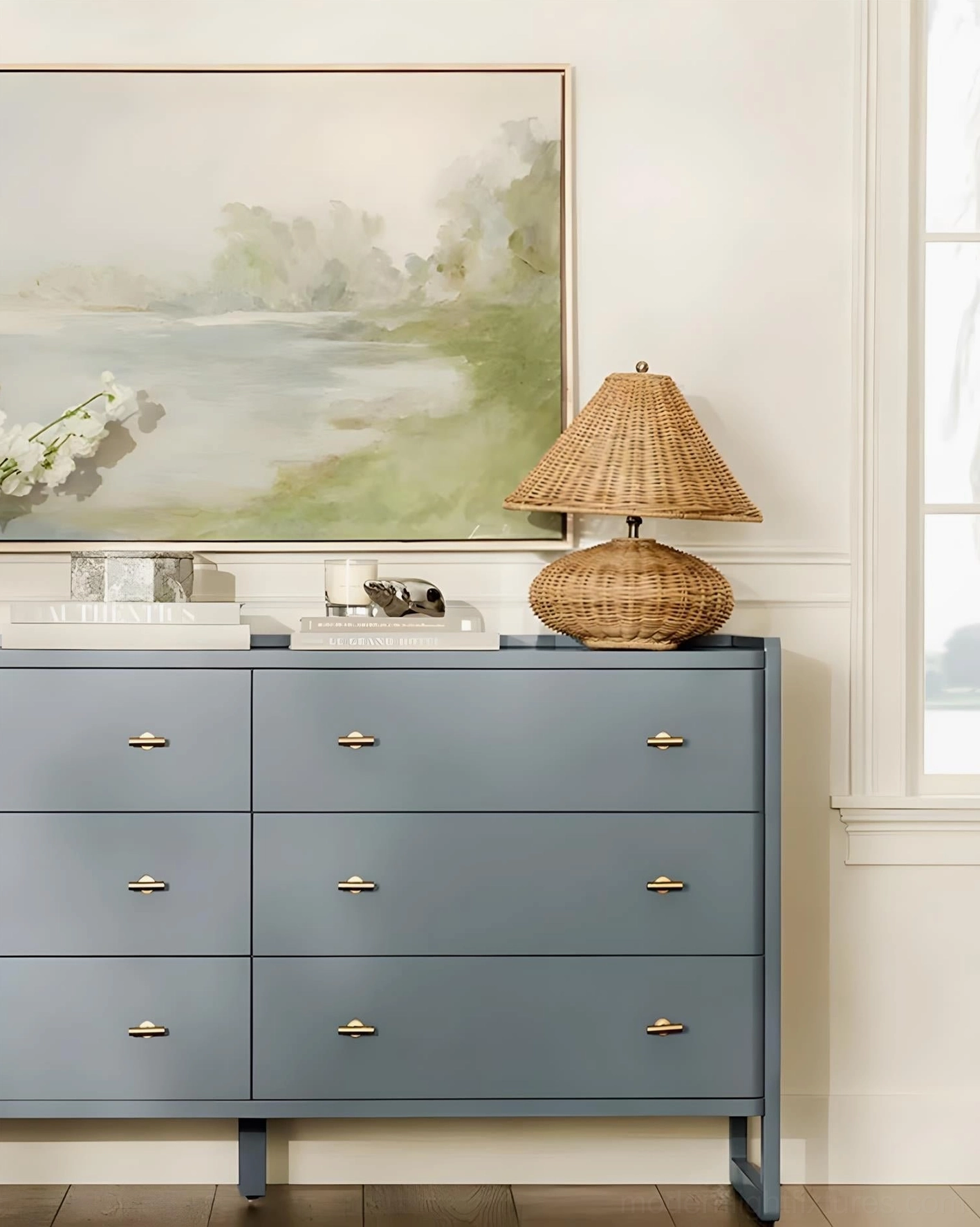 Rattan table lamp on blue bedroom dresser with landscape painting decor above, front side view showcasing coastal bohemian interior design