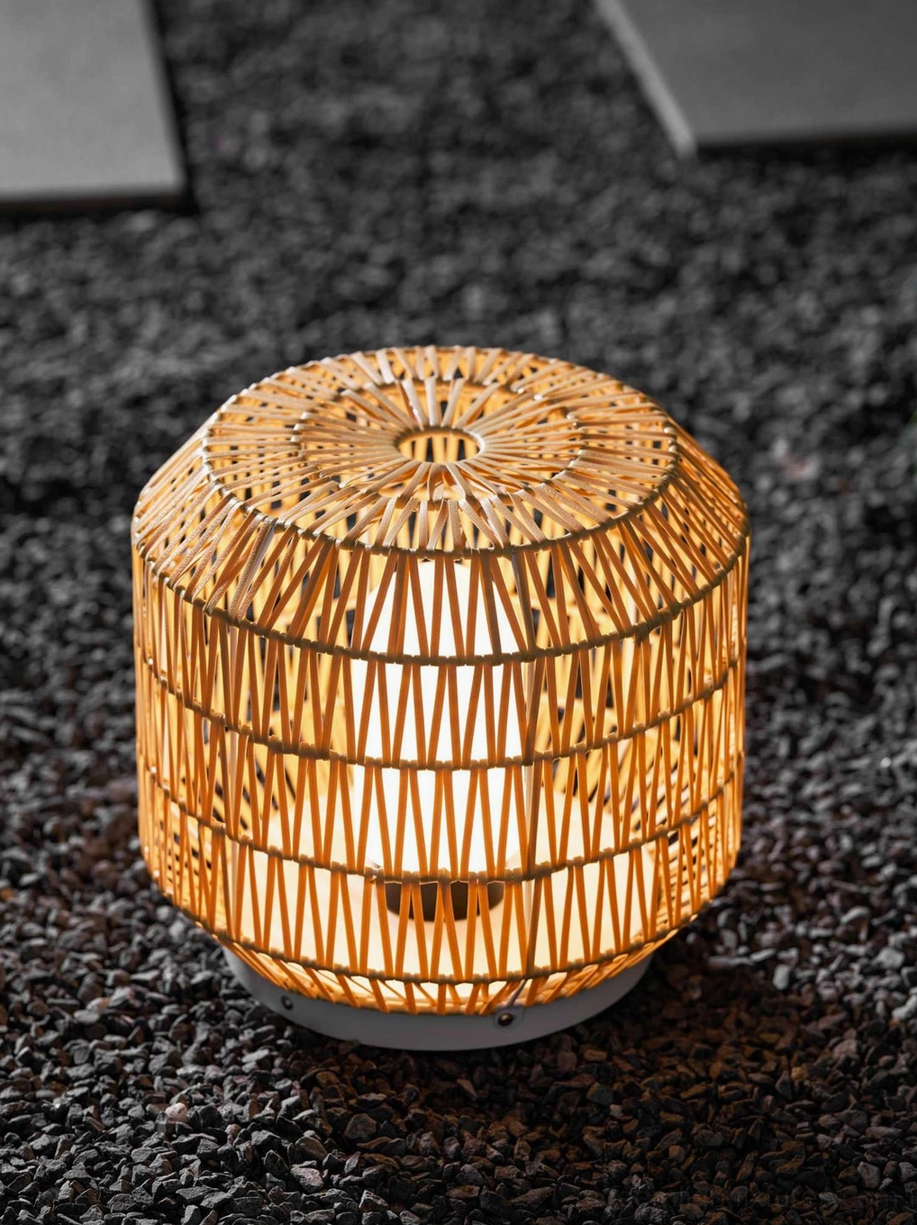 Close-up top-front view of woven rattan outdoor lantern lamp on gravel patio with warm illumination