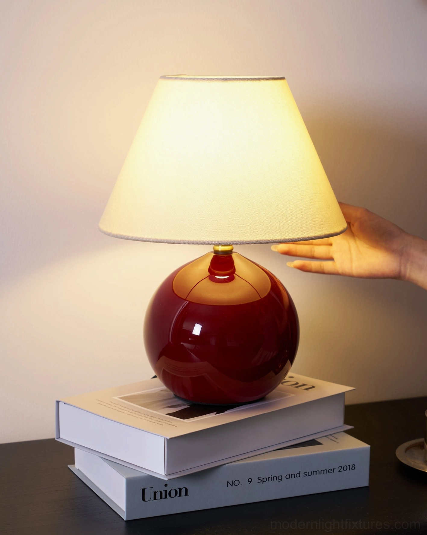 Hand presenting red sphere table lamp, showing scale and proportions on dark wood table