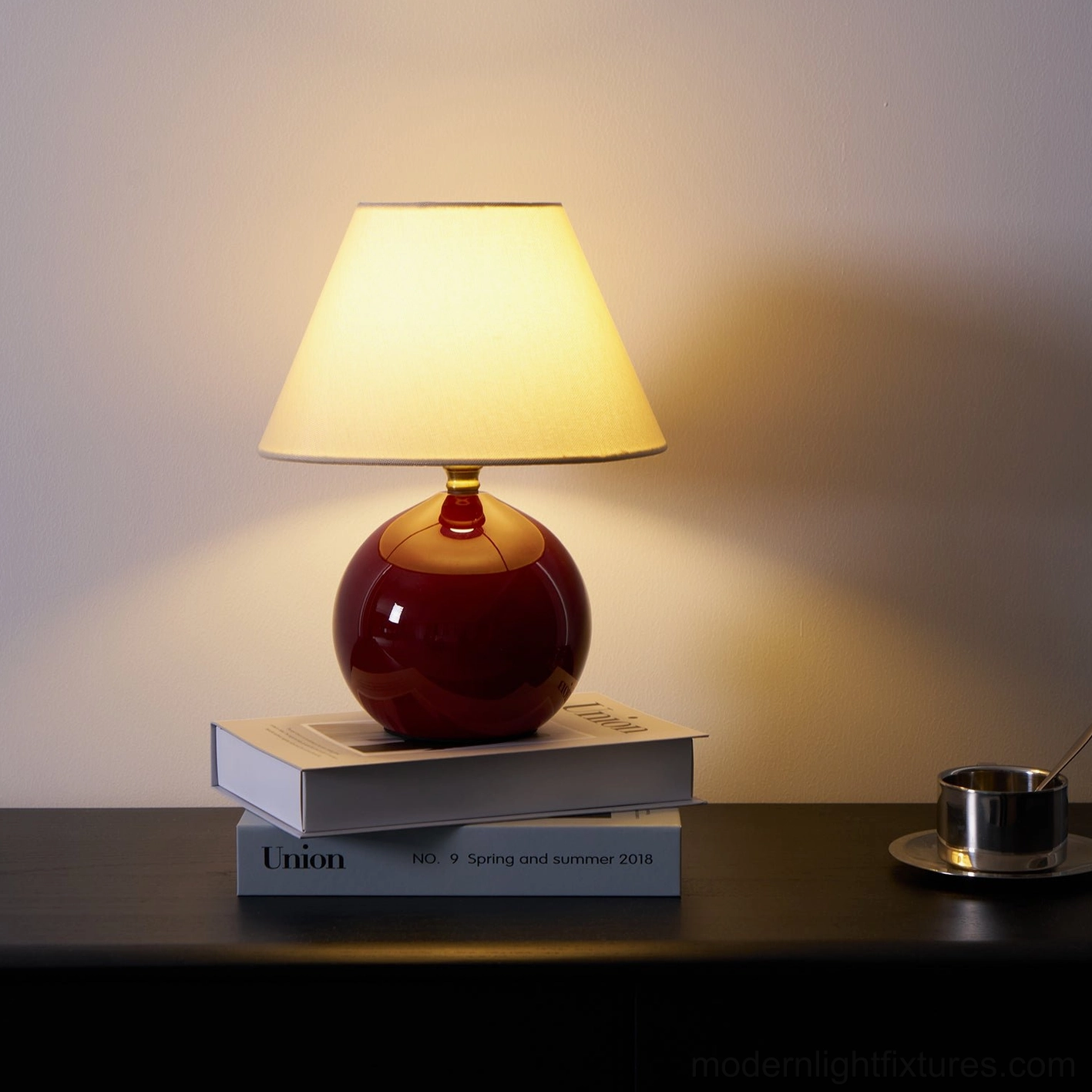 Side-front view of illuminated red table lamp on dark wood desk with stacked books, clear lampshade view