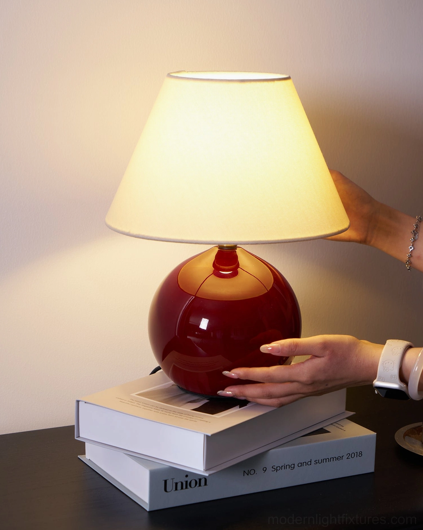 Hands adjusting red spherical table lamp base on dark wood surface, low angle product view