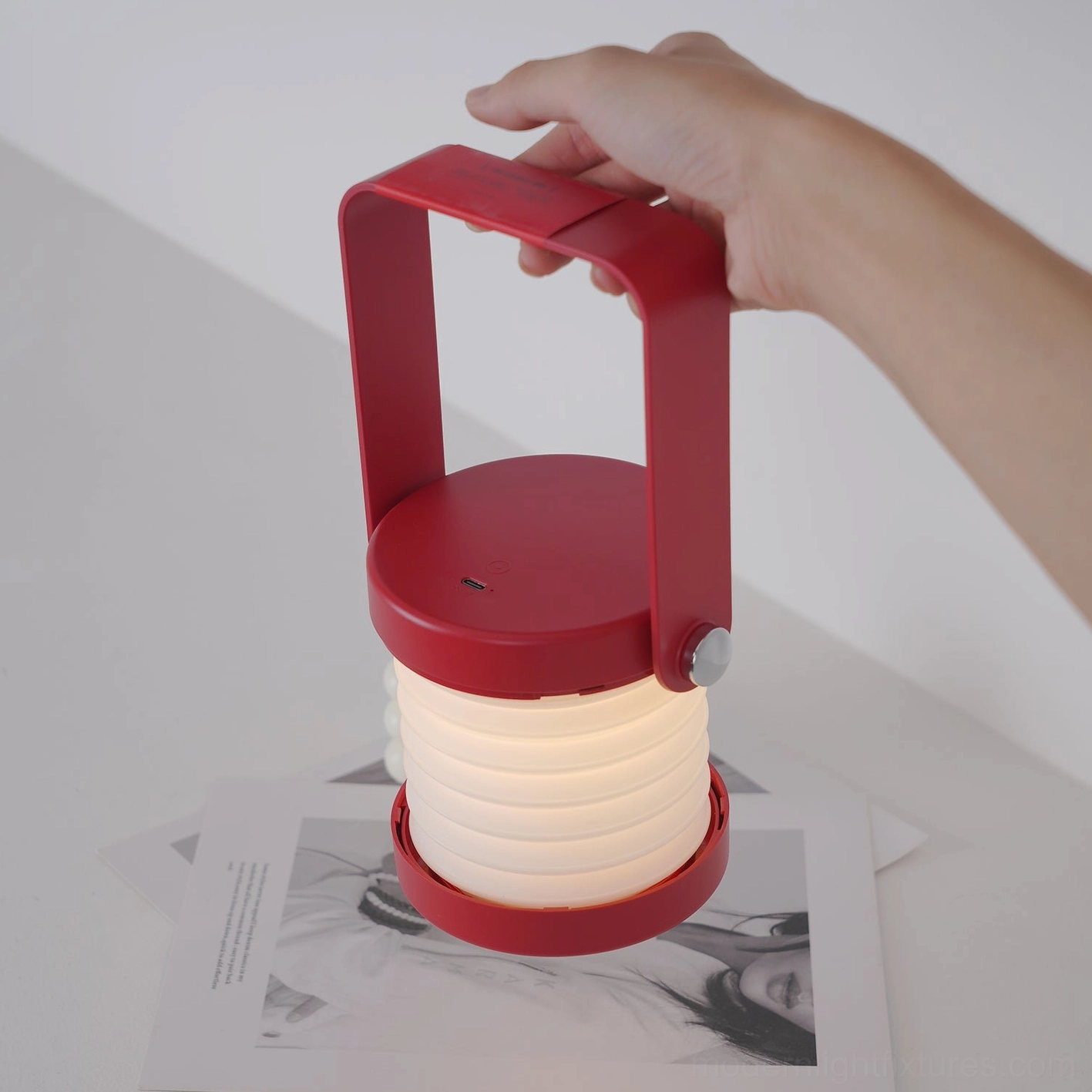 Red Terna Portable Lantern Lamp - Side oblique view with hand holding carrying handle, illuminated accordion lamp shade against white studio background