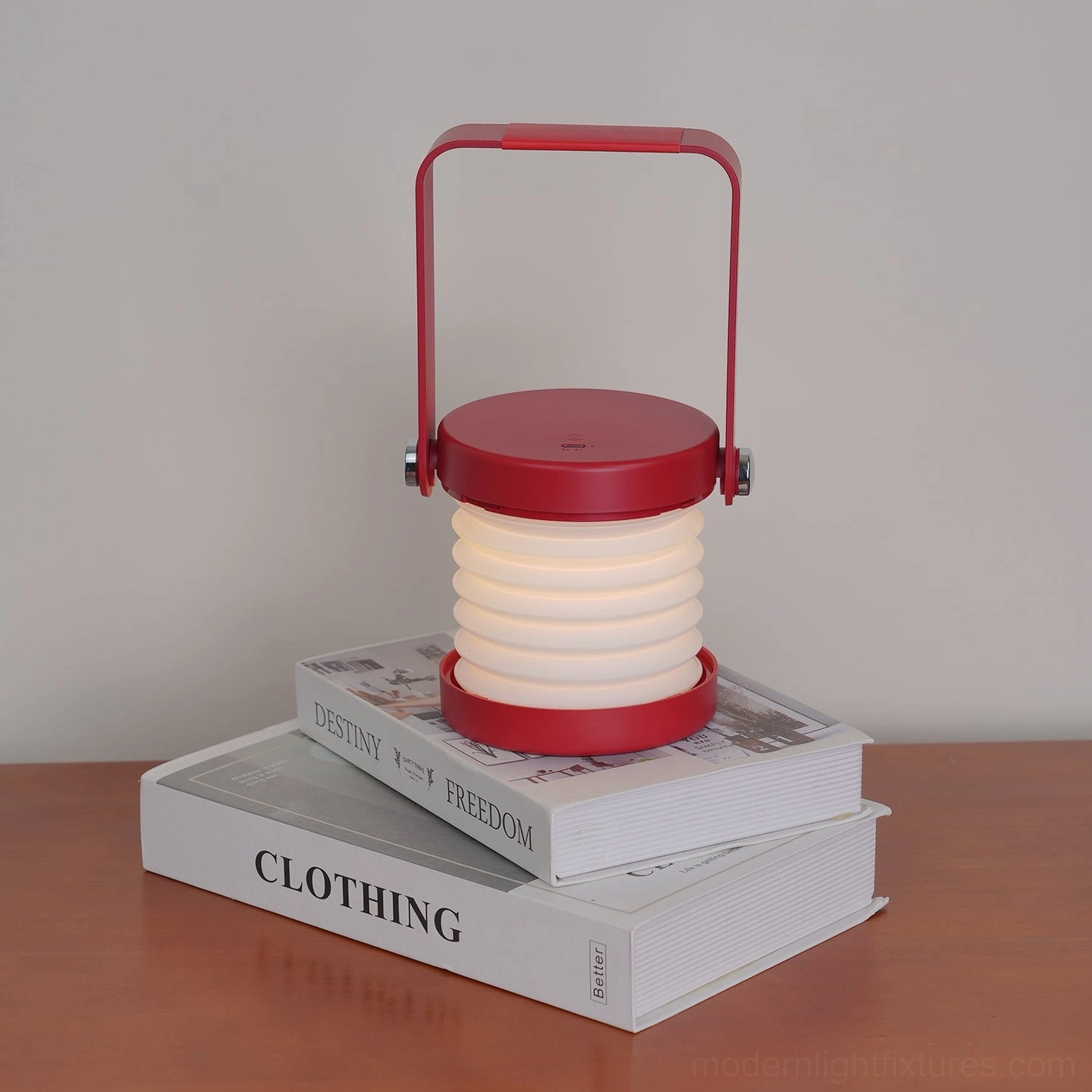 Red Terna Portable Lantern Lamp - Frontal view on wooden home desk stacked with books showing metal frame and white accordion shade