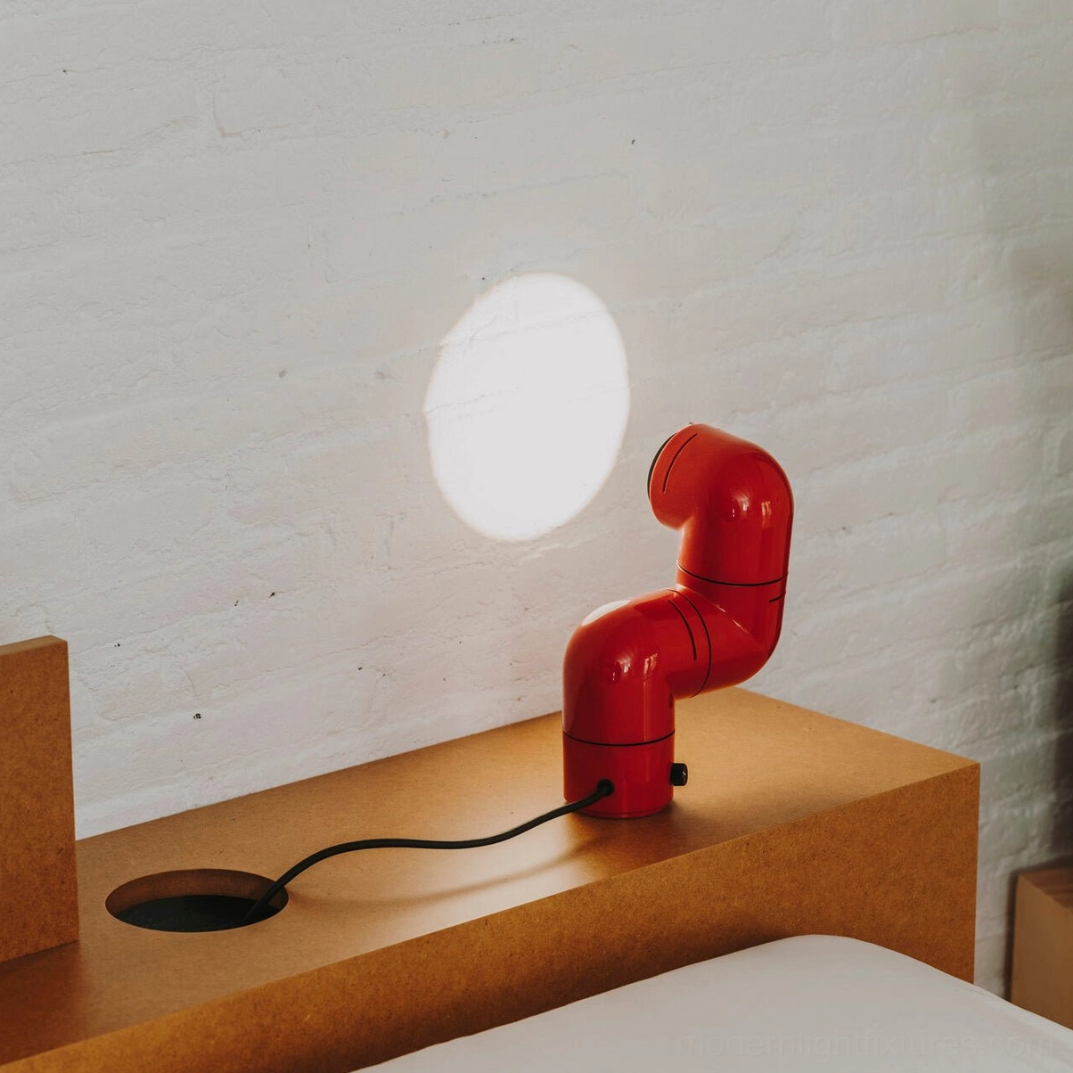 Red tubular table lamp on bedroom shelf projecting circular light spot on wall