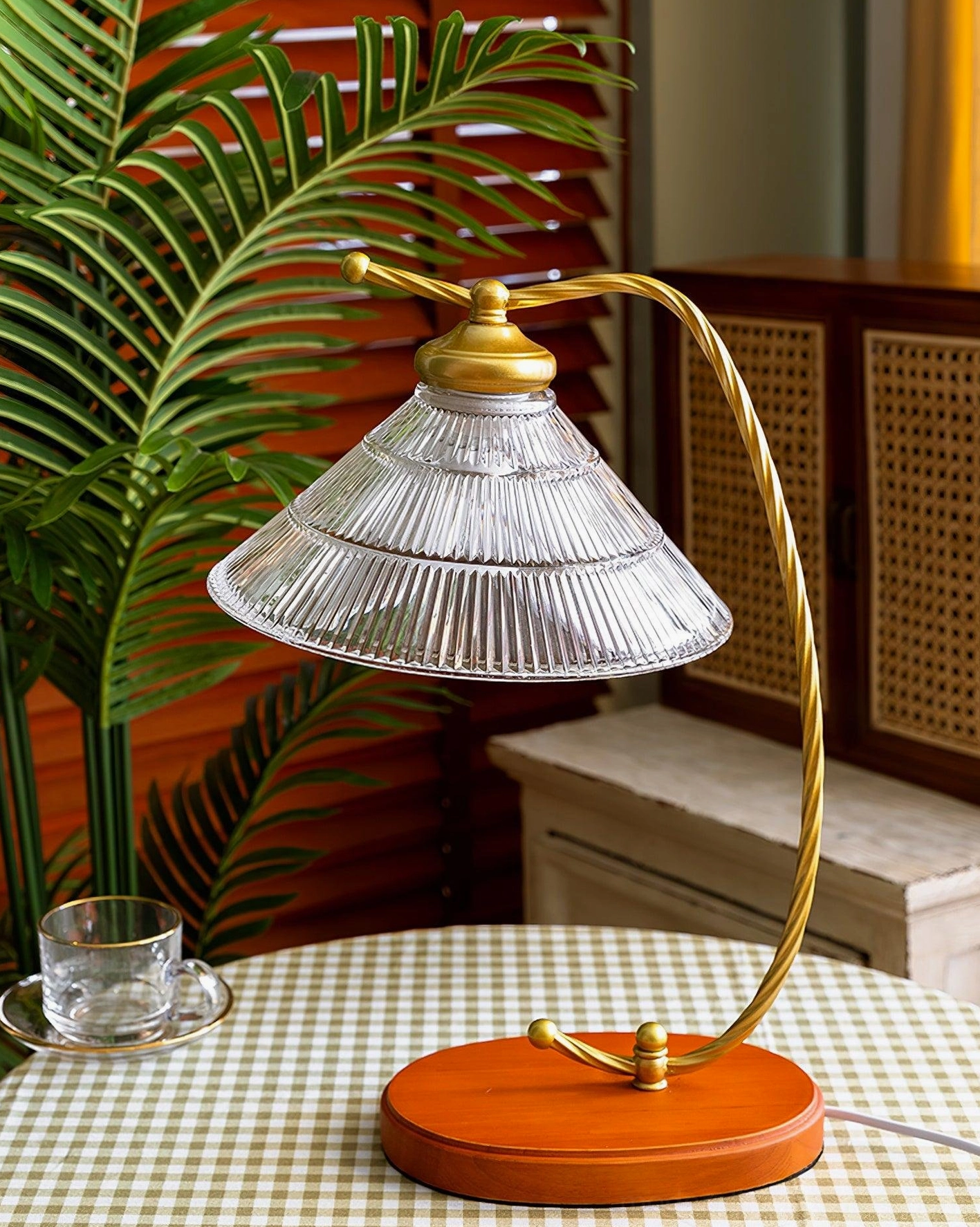 Mid-century arc table lamp with brass metal arm and terracotta base displayed on gingham tabletop with palm plant backdrop