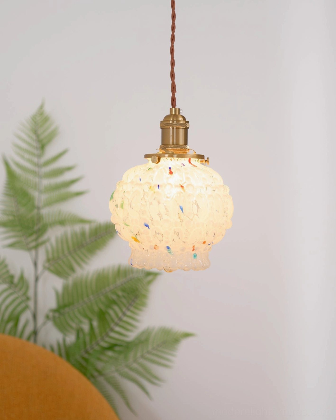 Lit retro brass pendant lamp with frosted hand-blown glass shade, colorful speckles, indoor fern plant corner, warm lighting