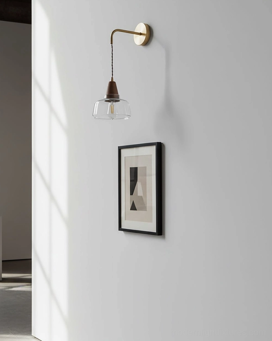 Minimalist plug-in wall sconce on residential hallway wall - slim brass arm with clear glass shade near framed art
