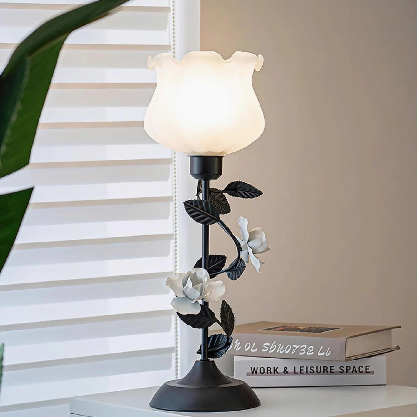Roselith rose table lamp three-quarter side view with warm glowing light, black metal stem with white floral accents in home interior setting