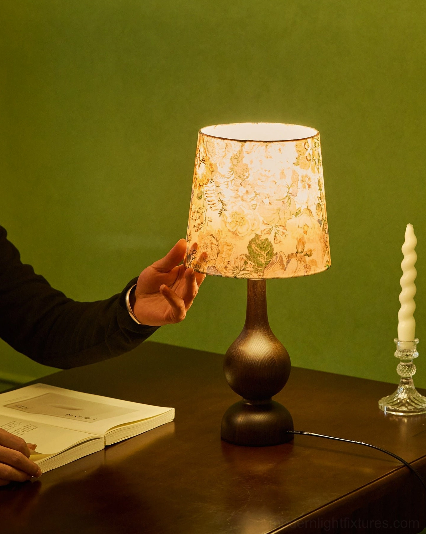 Vintage ash wood table lamp with floral print shade on wooden desk with green wall, side profile with hand adjusting
