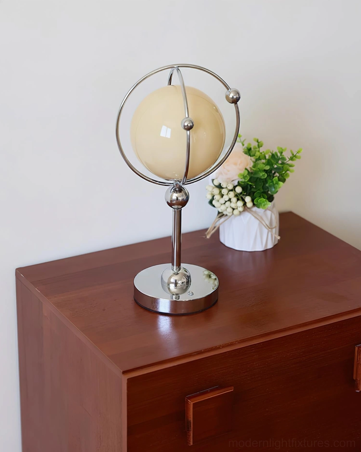 Saturn Hoop Table Lamp displayed on dark wood cabinet with potted flower arrangement and botanical styling