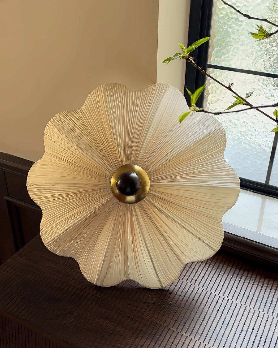 Seralyn pleated ceiling light displayed on wooden tabletop near window showing flower-shaped beige fabric shade with dark metal central fixture and natural décor