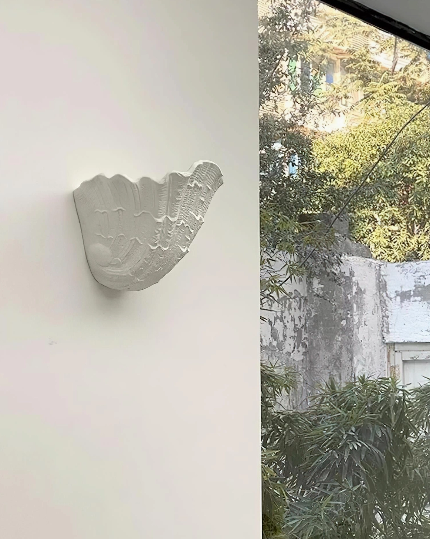 Coastal outdoor patio wall mounted shell sconce with matte white plaster finish and natural greenery backdrop