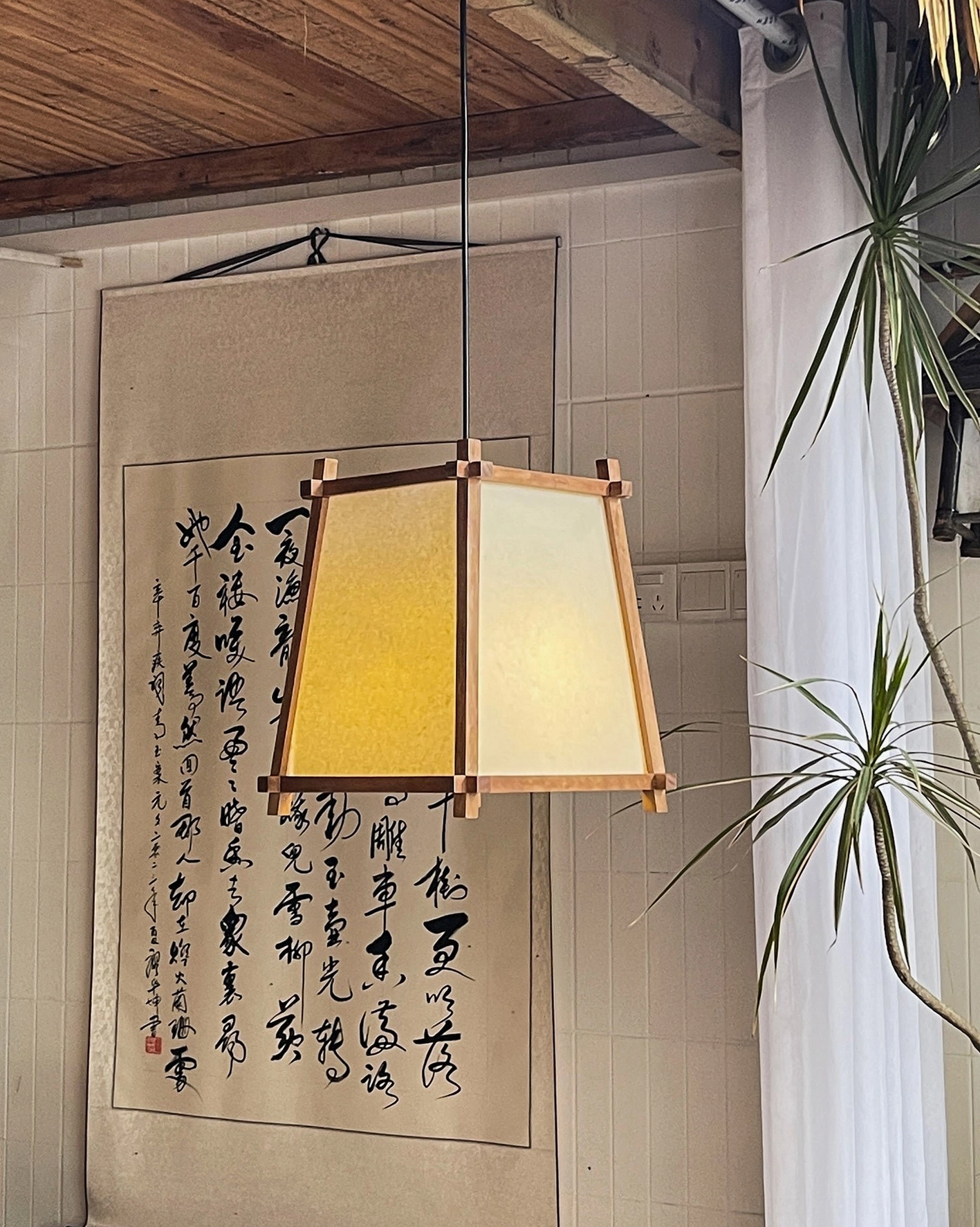 Shizuka wood pendant light three-quarter side view with calligraphy art and tropical plants in background
