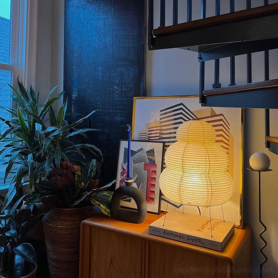 Warm glowing tiered paper lamp beside stacked books and artwork, bohemian reading nook